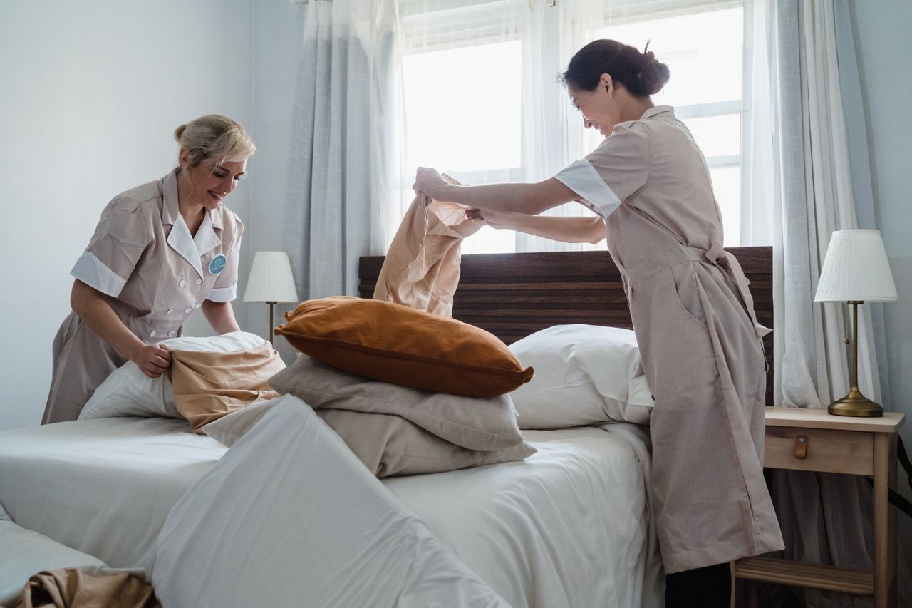 hero-img Two housekeepers in uniform fixing a hotel room bed with fresh linen and pillows.