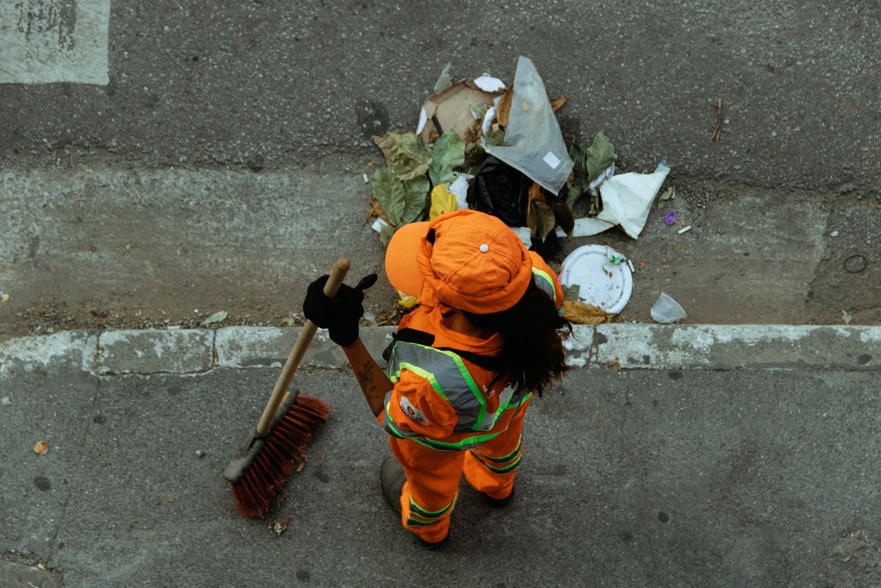 services-01 Top view of a street cleaner wearing orange uniform sweeping debris on a city sidewalk.